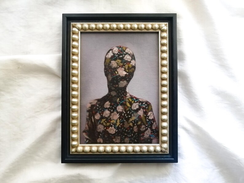 May include: Framed artwork featuring a figure with a floral pattern covering the head and torso. The frame is black with a gold-colored beaded inner border. The artwork is set against a light gray background.