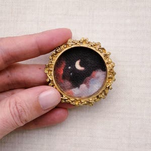 May include: A miniature painting in a round, ornate gold frame. The artwork depicts a night sky with a crescent moon, stars, and colorful clouds in shades of red, black, and white. The frame has a detailed, textured design.