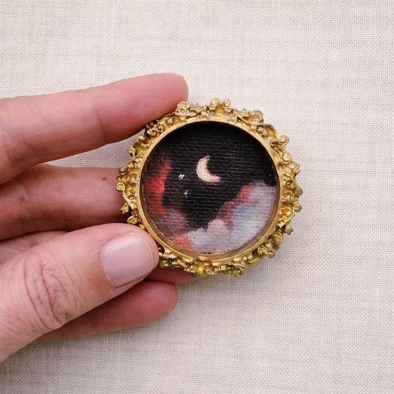 May include: A miniature painting in a round, ornate gold frame. The artwork depicts a night sky with a crescent moon, stars, and colorful clouds in shades of red, black, and white. The frame has a detailed, textured design.