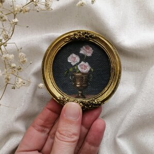 A small, round, gold-framed painting featuring a still life of pink roses in a gold vase against a dark background. The frame has ornate floral detailing. The painting is a miniature artwork.