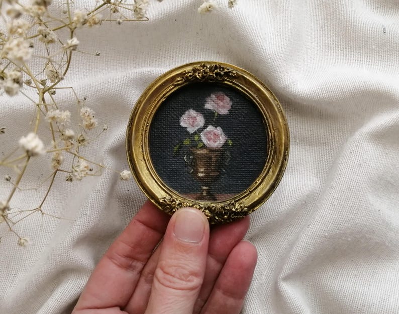A small, round, gold-framed painting featuring a still life of pink roses in a gold vase against a dark background. The frame has ornate floral detailing. The painting is a miniature artwork.