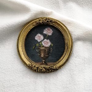 A small, round, antique gold-colored frame with a floral design. Inside the frame is a painting of pink roses in a gold vase against a dark blue background. The painting is a miniature still life.