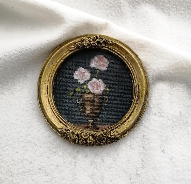 A small, round, antique gold-colored frame with a floral design. Inside the frame is a painting of pink roses in a gold vase against a dark blue background. The painting is a miniature still life.