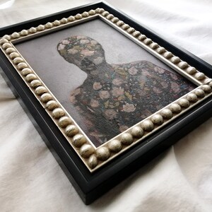 May include: A framed artwork featuring a figure with a floral pattern. The black frame has a decorative, beaded inner border. The artwork's subject is a person-shaped silhouette covered in a pattern of pink and white flowers.