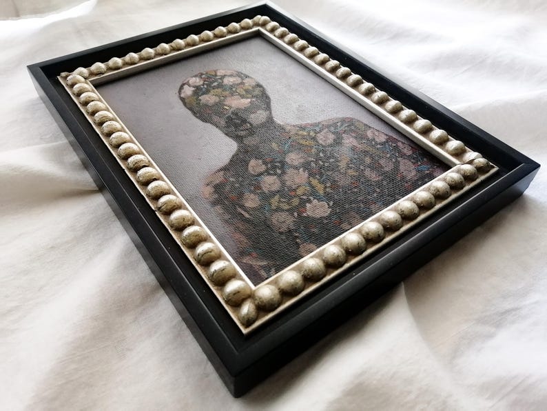 May include: A framed artwork featuring a figure with a floral pattern. The black frame has a decorative, beaded inner border. The artwork's subject is a person-shaped silhouette covered in a pattern of pink and white flowers.