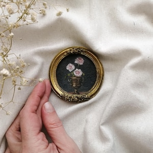 A tiny, round, gold-framed painting featuring a still life of pink roses in a gold vase against a dark background. The frame has ornate floral detailing. The painting is a miniature artwork.