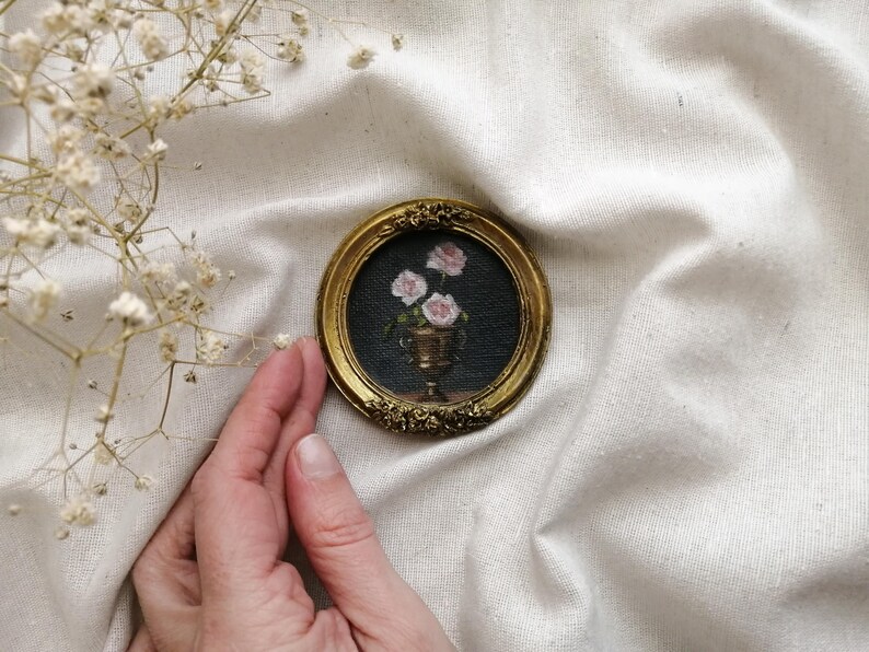A tiny, round, gold-framed painting featuring a still life of pink roses in a gold vase against a dark background. The frame has ornate floral detailing. The painting is a miniature artwork.