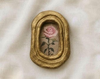 May include: An oval-shaped, textured gold frame containing a small, embroidered fabric insert. The embroidery features a single pink rose with green leaves and a stem, centered within the frame. The frame has a thick, uneven, and rustic border.