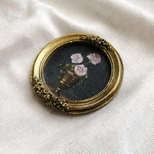 Op de afbeelding: Een kleine, ronde, antiek goudkleurige lijst met een miniatuurschilderij van roze rozen in een bronzen vaas tegen een donkere achtergrond. De lijst heeft sierlijke bloemendetails, wat een vintage esthetiek cre&euml;ert.
