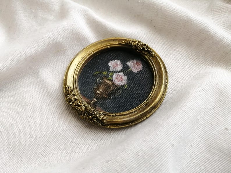 Op de afbeelding: Een kleine, ronde, antiek goudkleurige lijst met een miniatuurschilderij van roze rozen in een bronzen vaas tegen een donkere achtergrond. De lijst heeft sierlijke bloemendetails, wat een vintage esthetiek cre&euml;ert.