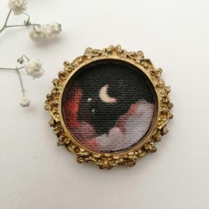 May include: A small, round painting of a night sky with a crescent moon and clouds, set in a gold-colored ornate frame. The artwork features a dark background with red, white, and gray clouds.