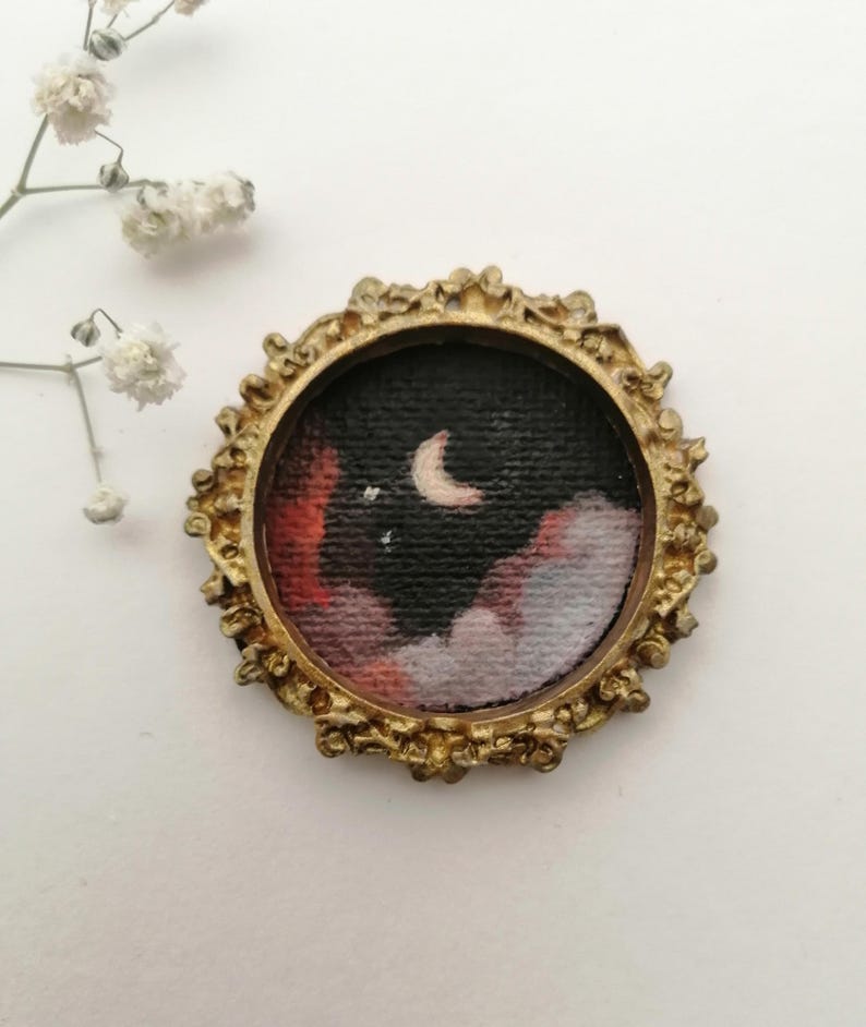 May include: A small, round painting of a night sky with a crescent moon and clouds, set in a gold-colored ornate frame. The artwork features a dark background with red, white, and gray clouds.