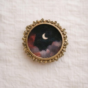 May include: A round, antique-style brooch with a miniature painting of a crescent moon in a dark sky with clouds. The painting is set in a gold-toned frame with a textured, beaded edge.
