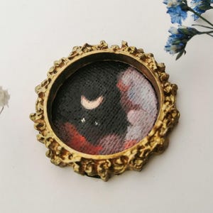 May include: A small, round painting in a gold-colored ornate frame. The artwork depicts a night sky with a crescent moon and stars, with red and white clouds. The frame has a detailed, textured design.