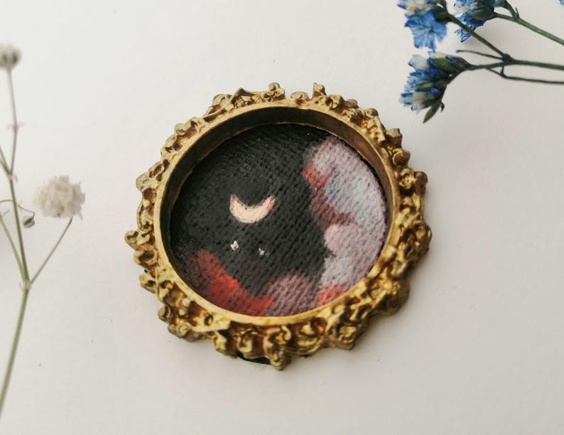 May include: A small, round painting in a gold-colored ornate frame. The artwork depicts a night sky with a crescent moon and stars, with red and white clouds. The frame has a detailed, textured design.