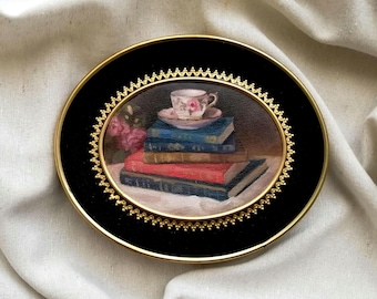 May include: An oval-shaped wall plaque featuring a miniature oil painting of a stack of four books topped with a floral teacup and saucer. The painting is encased in a black velvet border with a decorative gold-toned scalloped metal frame.