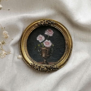 A miniature , round, gold-framed painting featuring a still life of pink roses in a gold vase against a dark background. The frame has ornate floral detailing. The painting is a miniature artwork.