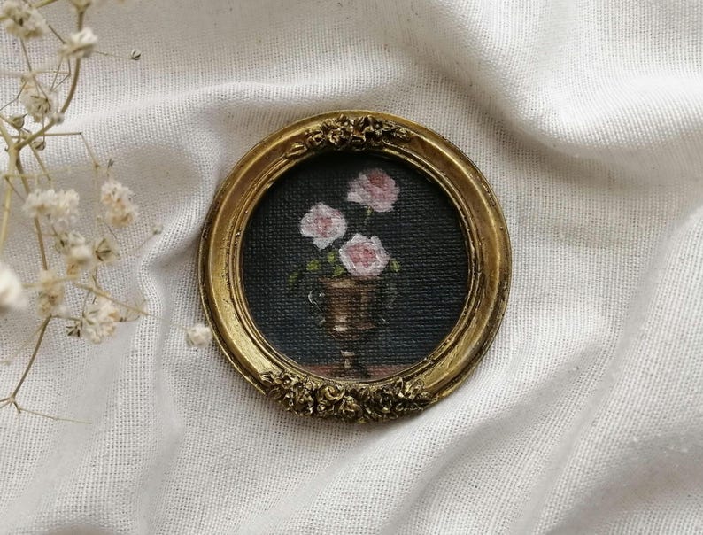 A miniature , round, gold-framed painting featuring a still life of pink roses in a gold vase against a dark background. The frame has ornate floral detailing. The painting is a miniature artwork.