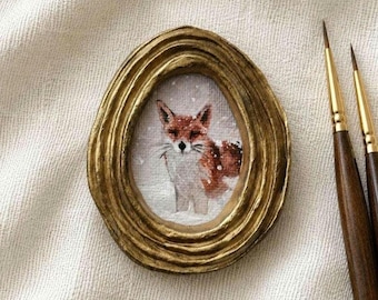 Puede incluir: Una pequeña pintura ovalada de un zorro en la nieve, dentro de un marco dorado metálico texturizado con bordes concéntricos. Dos pinceles finos con mangos de madera oscura y virolas doradas están situados a la derecha del marco.