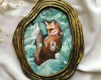 Puede incluir: Un marco dorado texturizado de forma ovalada que contiene una pintura de un zorro rojo acurrucado entre tela suave de color verde claro y pequeñas flores blancas. El zorro de pelaje marrón anaranjado está centrado en el marco ornamentado.