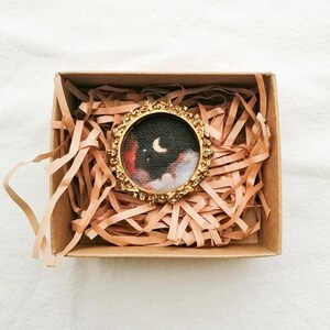 May include: A small, round painting of a night sky with a crescent moon and clouds, set in a gold-colored ornate frame. The artwork is presented inside a brown cardboard box filled with shredded paper for protection.