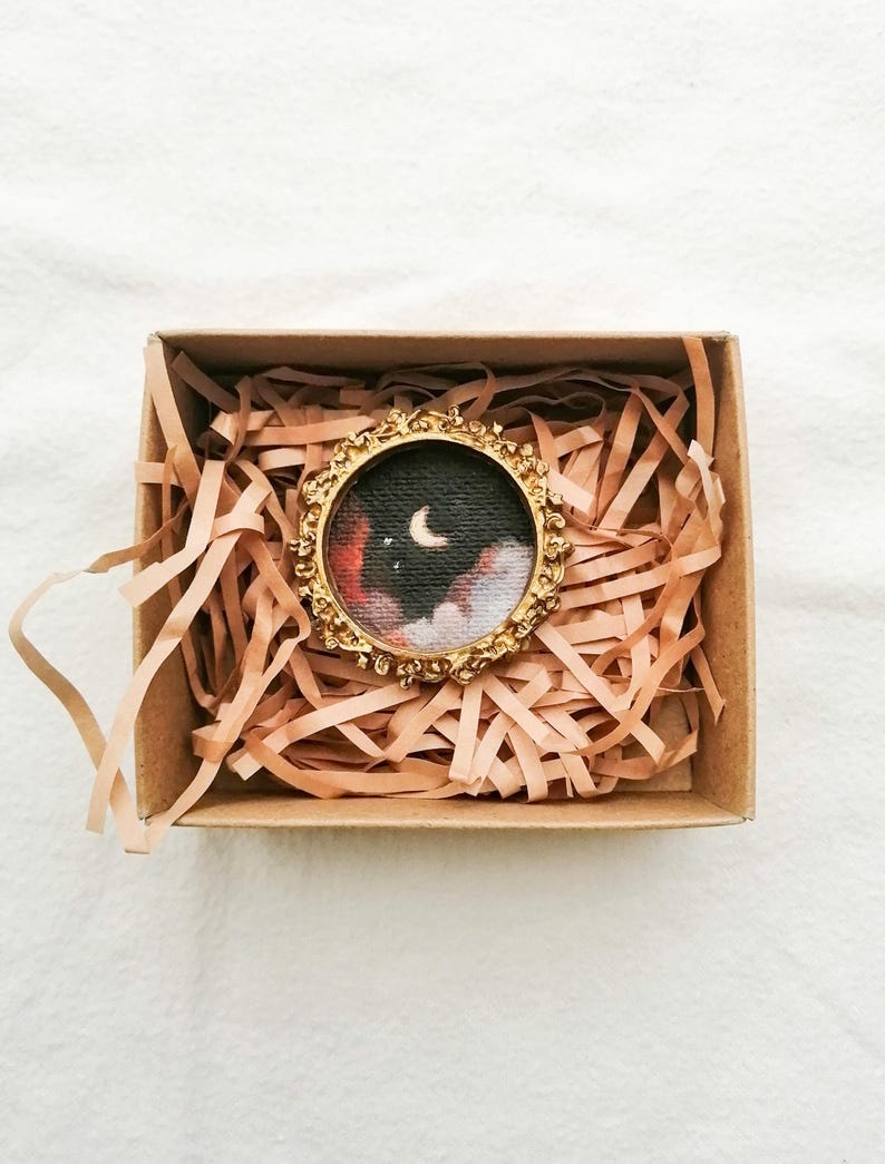 May include: A small, round painting of a night sky with a crescent moon and clouds, set in a gold-colored ornate frame. The artwork is presented inside a brown cardboard box filled with shredded paper for protection.