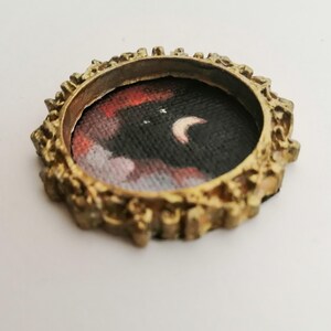May include: A small, round, antique gold-colored frame with a miniature painting of a night sky scene. The painting features a crescent moon and clouds in shades of red, white, and black.