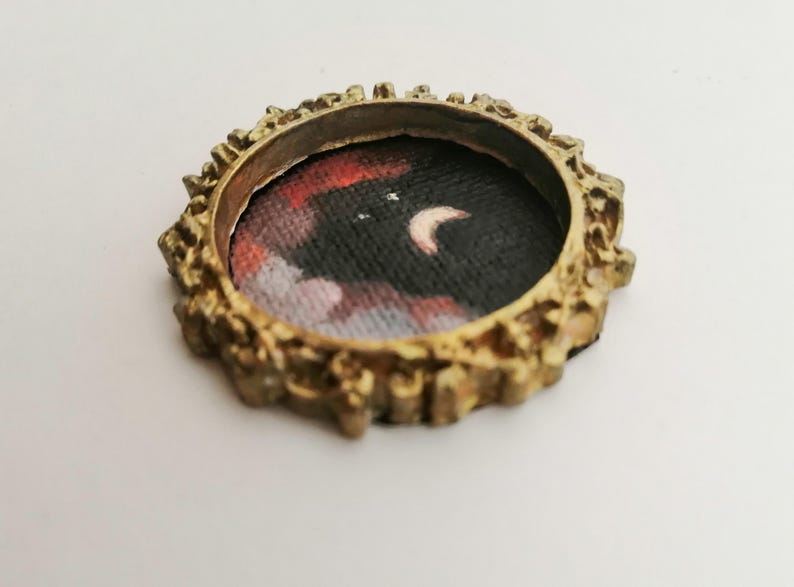 May include: A small, round, antique gold-colored frame with a miniature painting of a night sky scene. The painting features a crescent moon and clouds in shades of red, white, and black.