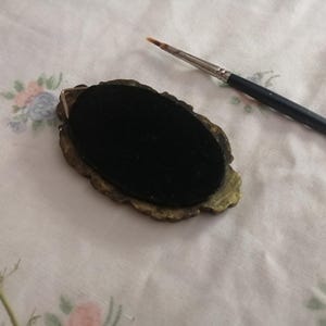 May include: An oval-shaped, antique-style jewelry component with a black velvet center and ornate gold-toned edges. A fine-tipped paintbrush rests nearby on a floral patterned fabric, suggesting artistic or crafting use.