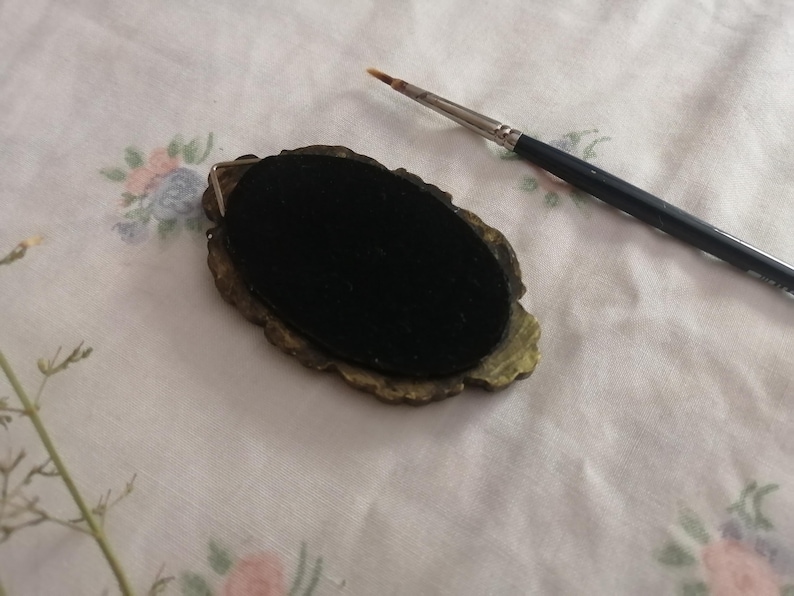 May include: An oval-shaped, antique-style jewelry component with a black velvet center and ornate gold-toned edges. A fine-tipped paintbrush rests nearby on a floral patterned fabric, suggesting artistic or crafting use.