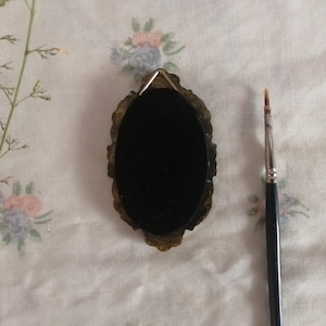 May include: An oval-shaped, dark-colored object with an ornate, gold-toned frame. A small paintbrush with a black handle is positioned to the right. The background is a white fabric with floral patterns and a sprig of dried plant.