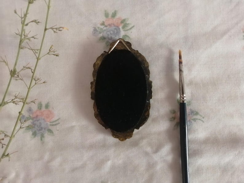 May include: An oval-shaped, dark-colored object with an ornate, gold-toned frame. A small paintbrush with a black handle is positioned to the right. The background is a white fabric with floral patterns and a sprig of dried plant.