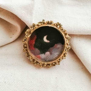 May include: A small, round, gold-colored brooch with an ornate, textured frame. The brooch features a miniature painting of a night sky with a crescent moon and colorful clouds in shades of red, pink, and black.