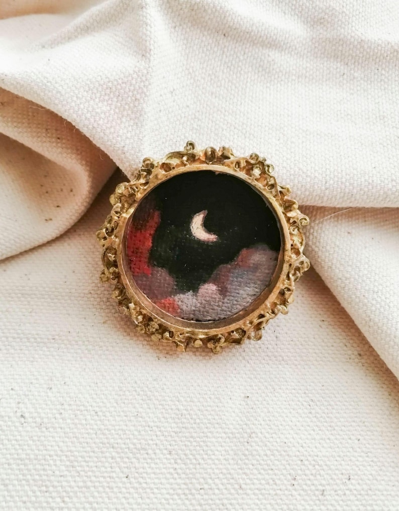 May include: A small, round, gold-colored brooch with an ornate, textured frame. The brooch features a miniature painting of a night sky with a crescent moon and colorful clouds in shades of red, pink, and black.