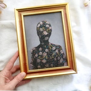 May include: A framed artwork featuring a figure with a floral pattern covering the head and torso. The frame is gold with a reddish-brown inner edge. The background is a light gray, and the artwork is held by a hand.