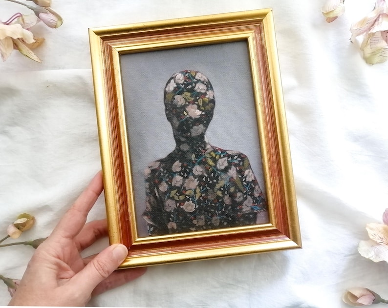 May include: A framed artwork featuring a figure with a floral pattern covering the head and torso. The frame is gold with a reddish-brown inner edge. The background is a light gray, and the artwork is held by a hand.