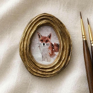 Op de afbeelding: Een ovaal miniatuurschilderij van een vos in de sneeuw, gevat in een getextureerde, goudkleurige lijst met concentrische ringdetails, geplaatst naast twee dunne penselen met donkere stelen, gouden hulzen en fijne, puntige haren.