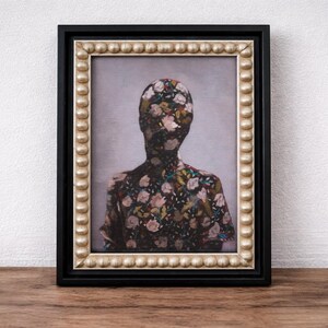 May include: Framed artwork featuring a figure with a floral pattern covering the head and upper body. The artwork is in a black frame with a gold beaded inner border. The background is a light gray color.