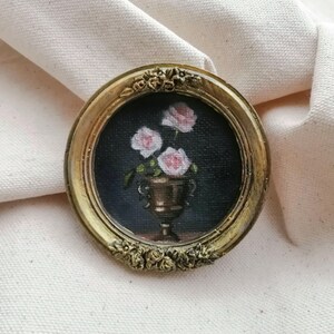 A small, round painting in a gold frame. The artwork depicts pink roses in a bronze vase against a dark Moody  background. The frame has ornate floral detailing.