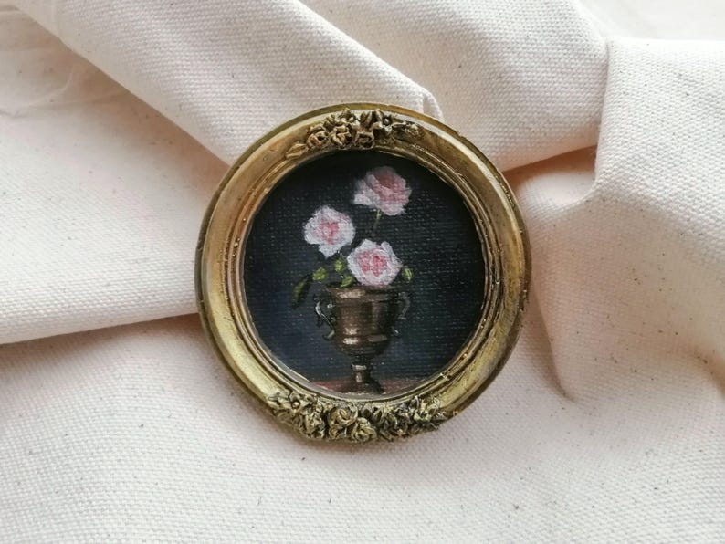 A small, round painting in a gold frame. The artwork depicts pink roses in a bronze vase against a dark Moody  background. The frame has ornate floral detailing.