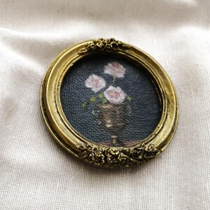 Op de afbeelding: Een kleine, ronde, antiek goudkleurige lijst met een miniatuurschilderij van roze rozen in een vaas. De schildering heeft een donkerblauwe achtergrond. De lijst heeft sierlijke bloemendetails.
