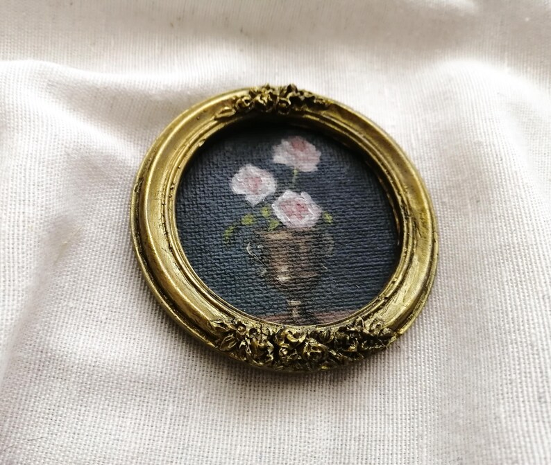 Op de afbeelding: Een kleine, ronde, antiek goudkleurige lijst met een miniatuurschilderij van roze rozen in een vaas. De schildering heeft een donkerblauwe achtergrond. De lijst heeft sierlijke bloemendetails.