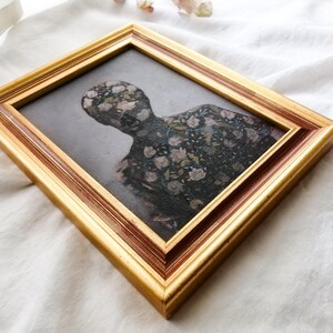 May include: A framed artwork featuring a figure with a floral pattern. The artwork is set within a gold-colored frame with a dark brown inner edge. The figure's form is filled with a pattern of flowers in shades of pink, white, and green.