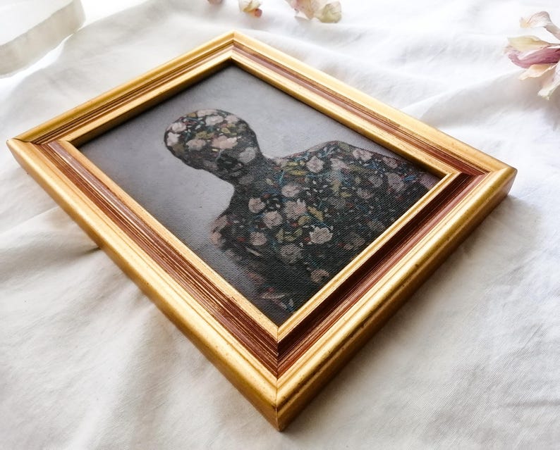 May include: A framed artwork featuring a figure with a floral pattern. The artwork is set within a gold-colored frame with a dark brown inner edge. The figure's form is filled with a pattern of flowers in shades of pink, white, and green.