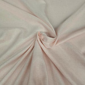 May include: A close-up shot of a light pink fabric with a subtle woven texture. The fabric is gathered and folded, creating soft shadows and highlights. The overall appearance is smooth and delicate, suggesting a lightweight material.