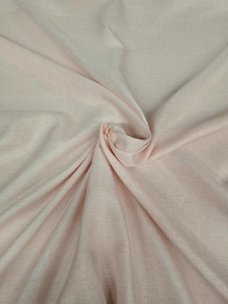 May include: A close-up shot of a light pink fabric with a subtle woven texture. The fabric is gathered and folded, creating soft shadows and highlights. The overall appearance is smooth and delicate, suggesting a lightweight material.