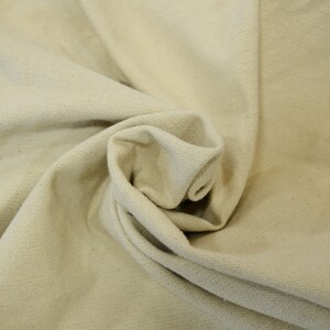 May include: Close-up of a natural off-white canvas fabric with a textured weave. The fabric is folded and draped, showing its soft and pliable texture.
