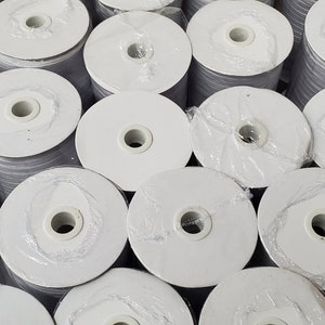 May include: A close-up of a box filled with spools of white elastic. The spools are wrapped in clear plastic and have a white center.