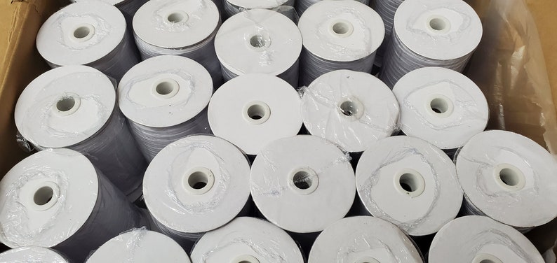 May include: A close-up of a box filled with spools of white elastic. The spools are wrapped in clear plastic and have a white center.