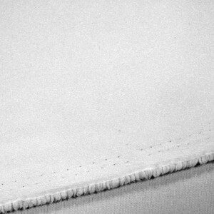 以下が含まれることがあります： テクスチャ加工された表面を持つ白い生地のクローズアップ。端はほつれており、端の近くにステッチの線が見えます。生地は、おそらく綿またはリネンである軽い素材のようです。